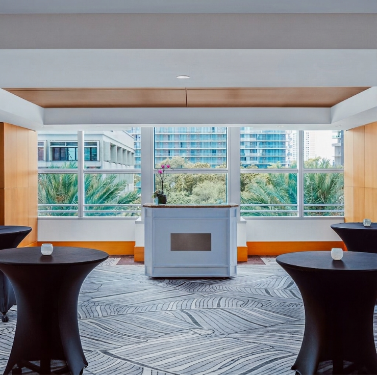 Modern meeting space with tall windows, a small bar or counter, round cocktail tables with black covers, and a view of trees and buildings outside.