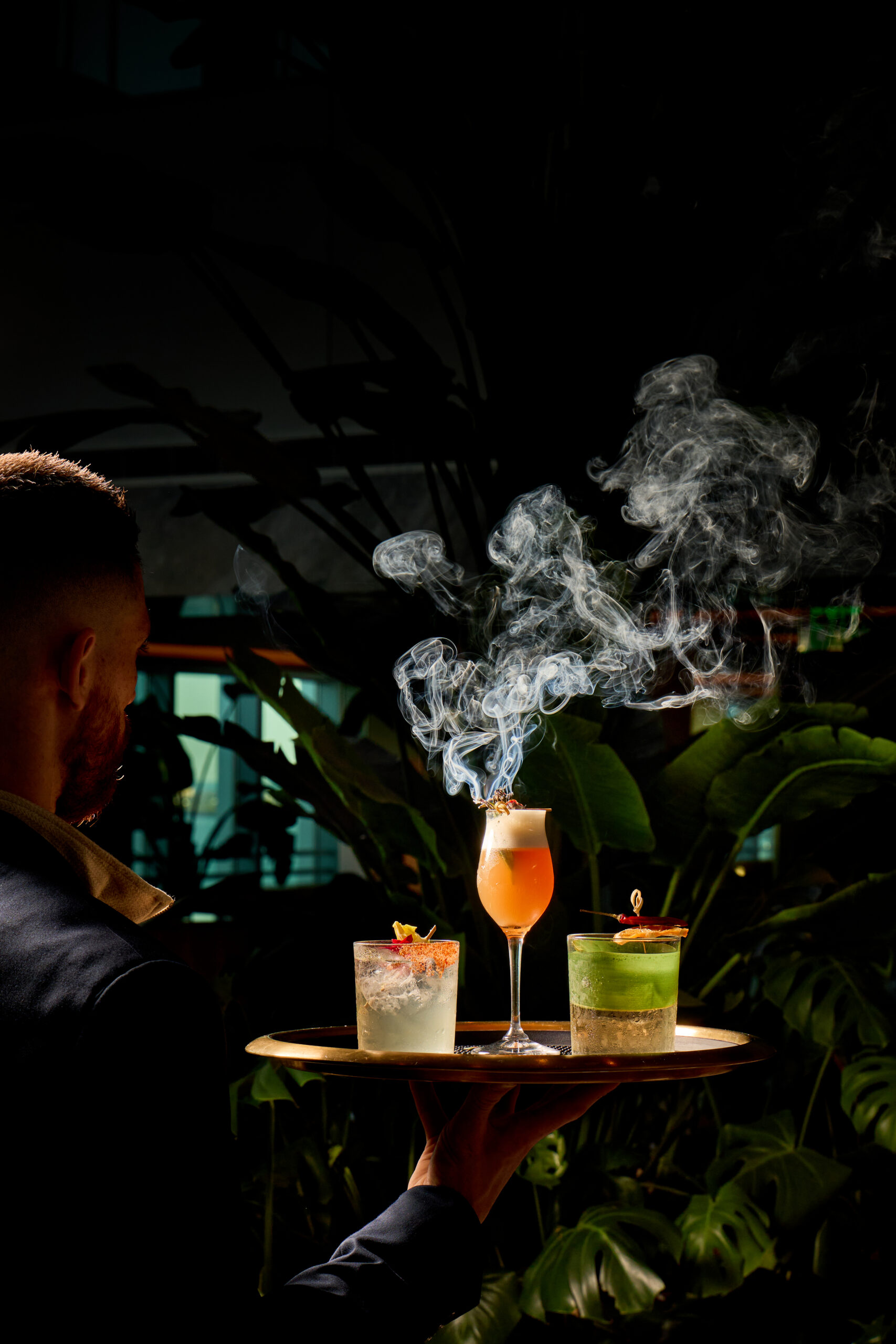 A person holds a tray with three cocktails, one with dramatic smoke rising, set against a background of lush green plants.
