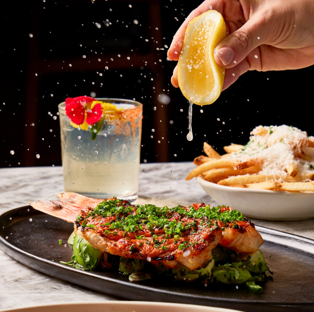 A hand squeezes a lemon over grilled shrimp on a platter, with a drink and a bowl of fries in the background.
