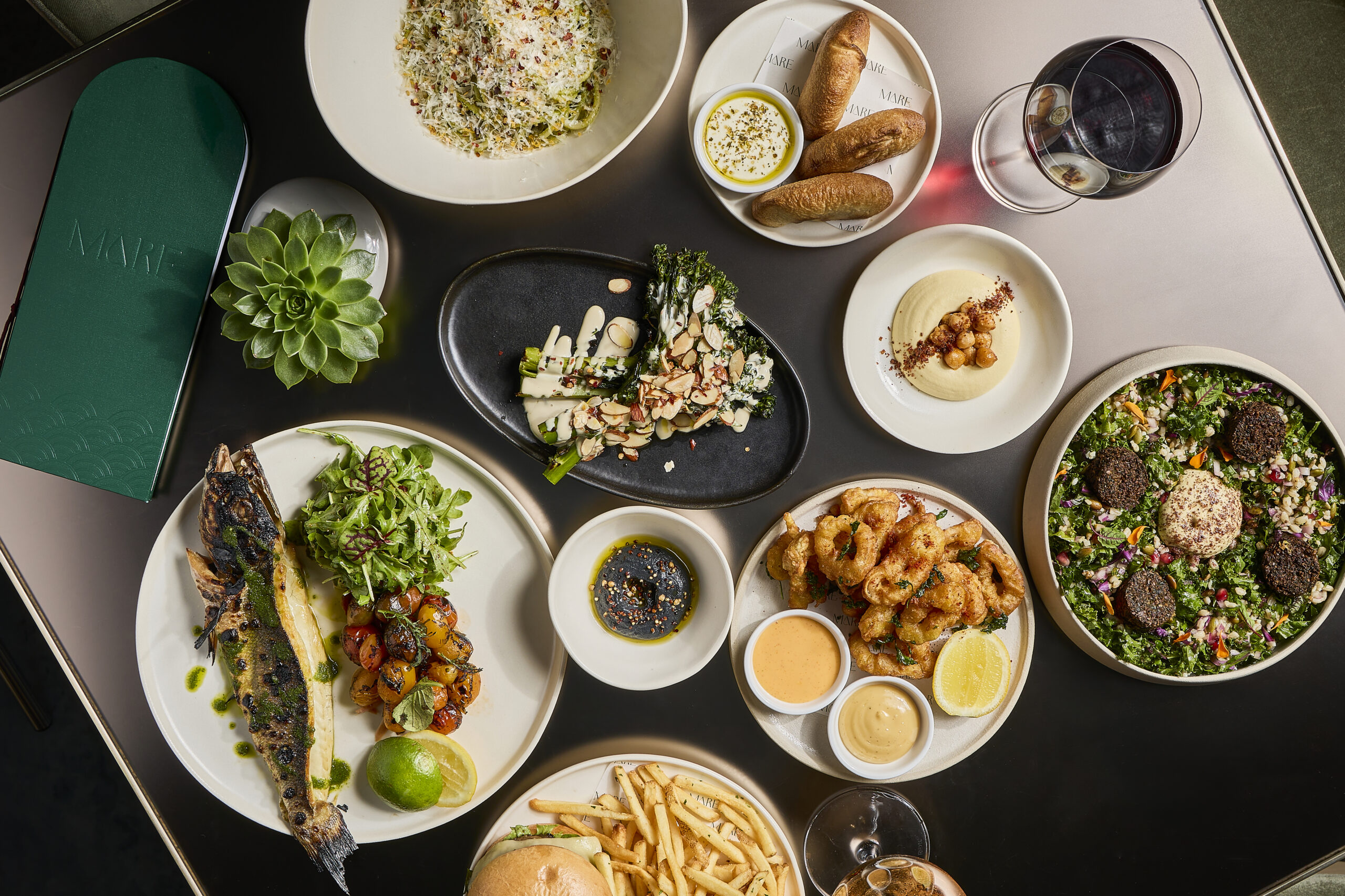 Overhead view of a table with plates of grilled fish, salad, shrimp, bread, sauces, fries, wine, and a green menu next to a small succulent plant.