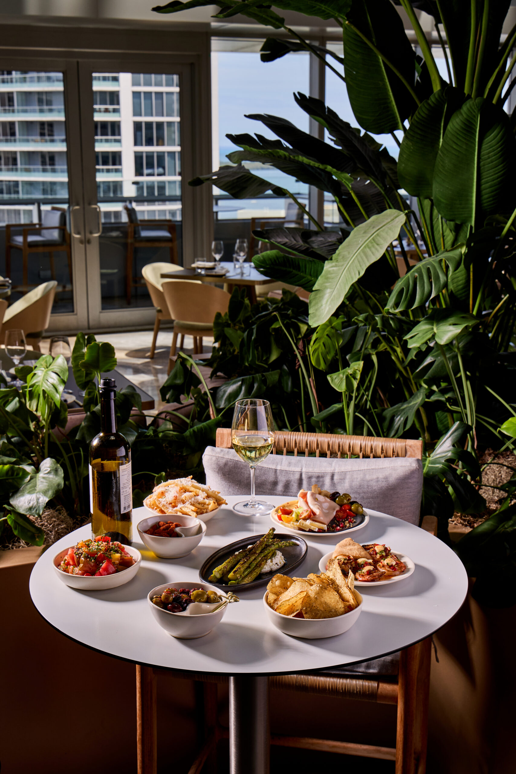 A table with wine, bread, chips, dips, vegetables, and appetizers, set near lush plants and large windows overlooking tall buildings.