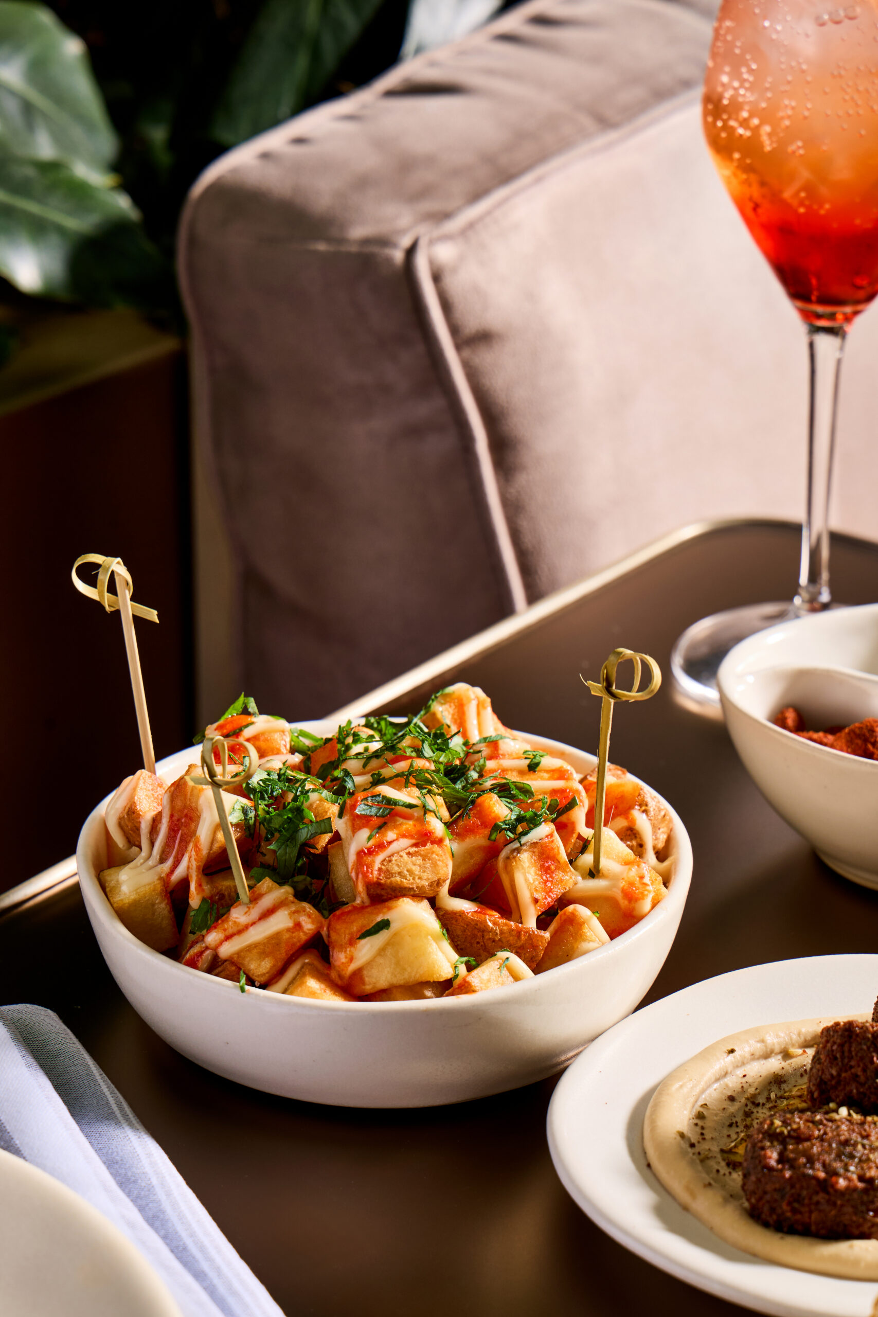 A bowl of patatas bravas topped with sauce and herbs sits on a table beside a plate of hummus and a glass of red-orange cocktail.