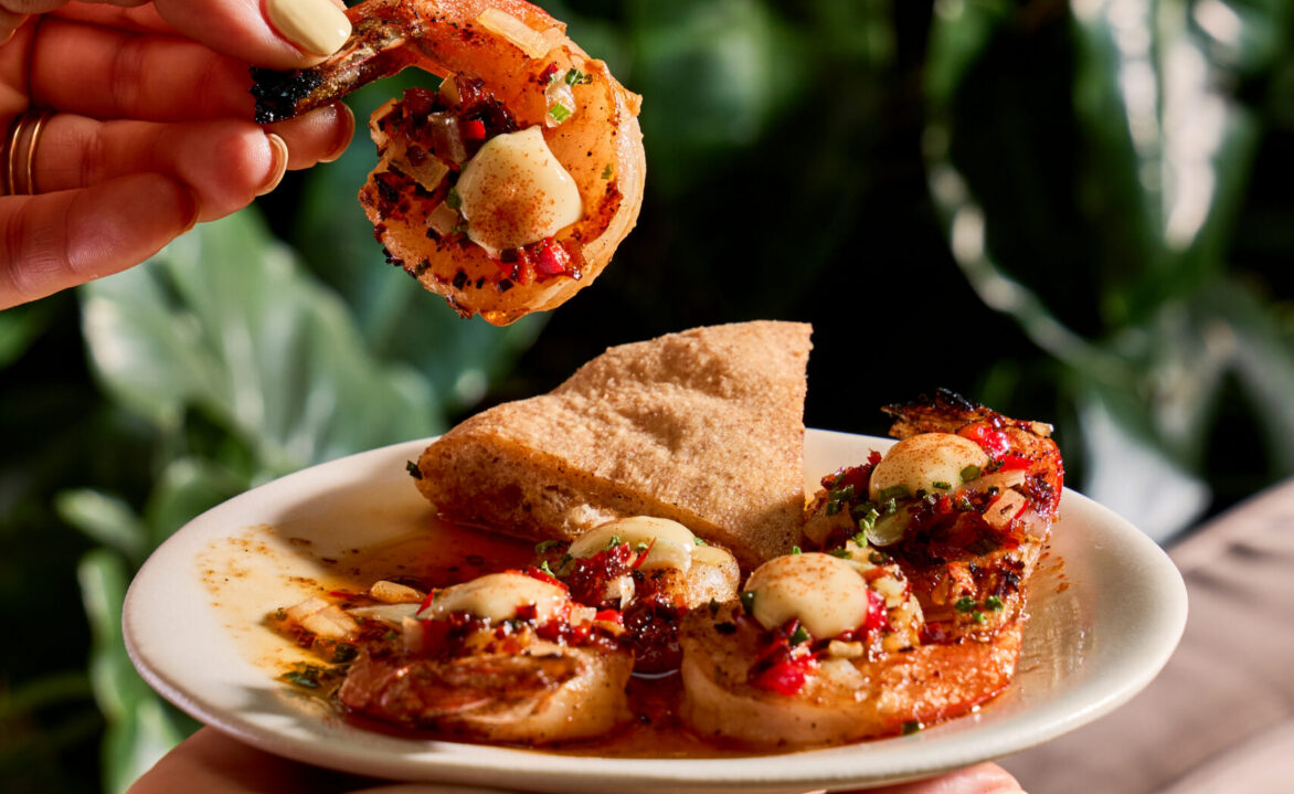 A hand holds a shrimp above a plate with shrimp, flatbread, and herbs, set against a background of green leafy plants.
