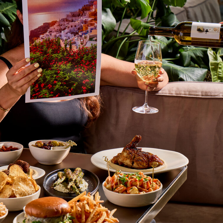 A person holds a photo of a scenic coastal town and a glass of white wine, sitting at a table filled with various dishes, including fries and pasta.