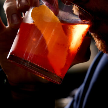 A person sipping an orange-colored cocktail with ice and an orange peel garnish from a glass, lit dramatically from the side.