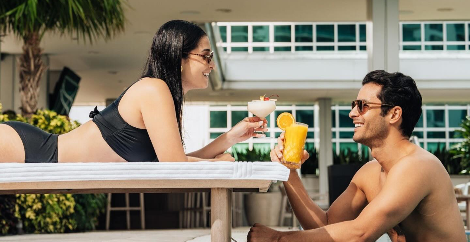 A woman lounges by a pool holding a cocktail, smiling at a man sitting in the water holding an orange drink.