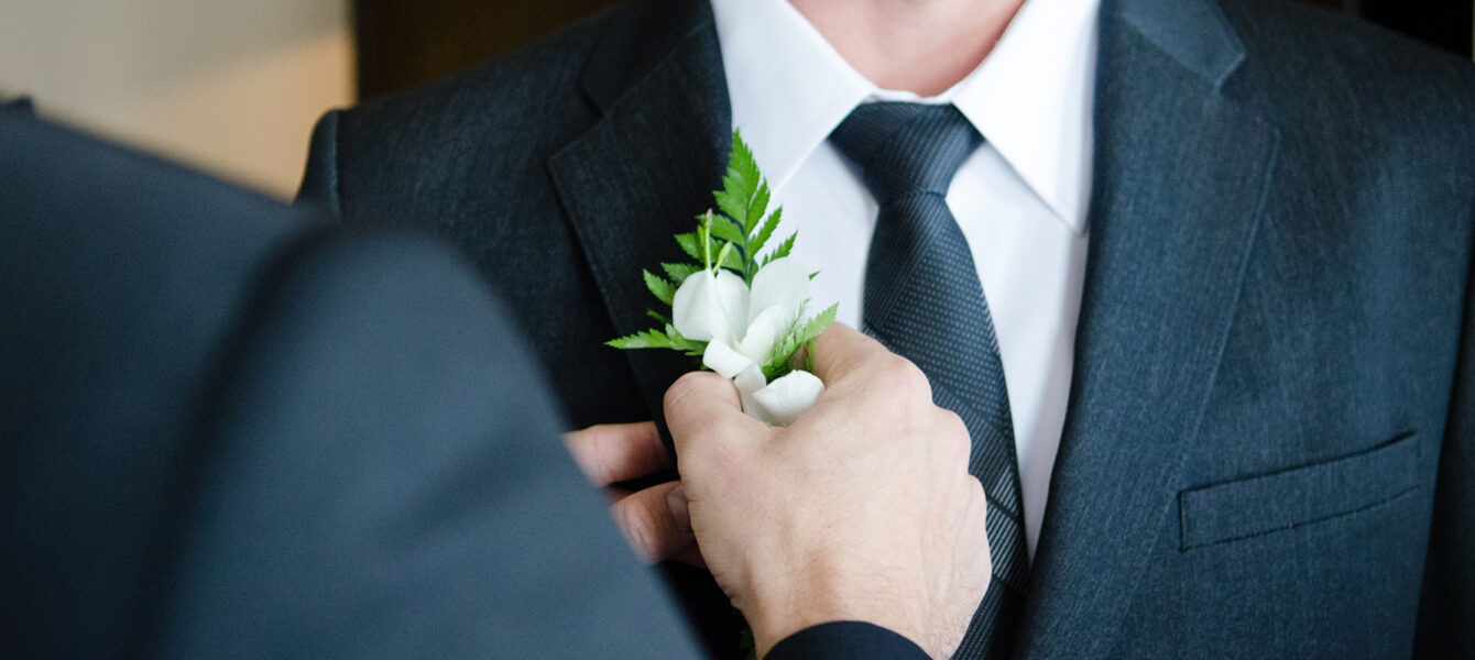 A person pins a white boutonniere with green leaves to the lapel of a man wearing a dark suit, white shirt, and striped tie.