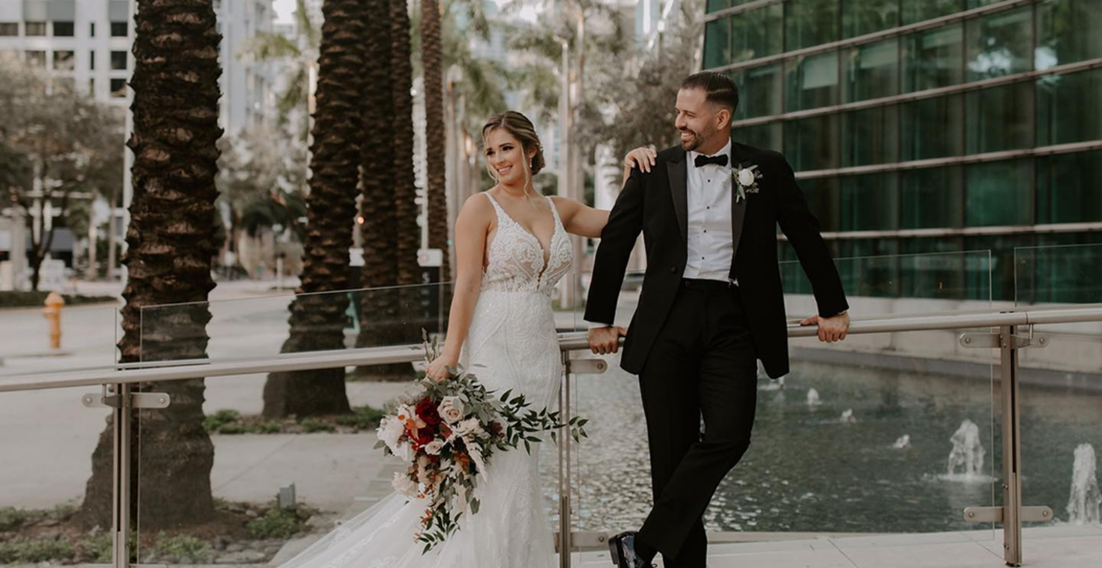 Bride in white gown and groom in black tuxedo pose by a fountain, holding a bouquet, with palm trees and city buildings in the background.