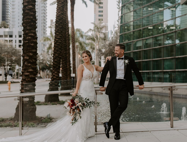 Bride in white gown and groom in black tuxedo pose by a fountain, holding a bouquet, with palm trees and city buildings in the background.