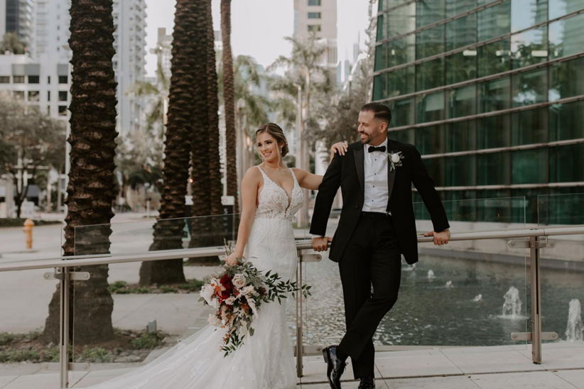 Bride in white gown and groom in black tuxedo pose by a fountain, holding a bouquet, with palm trees and city buildings in the background.