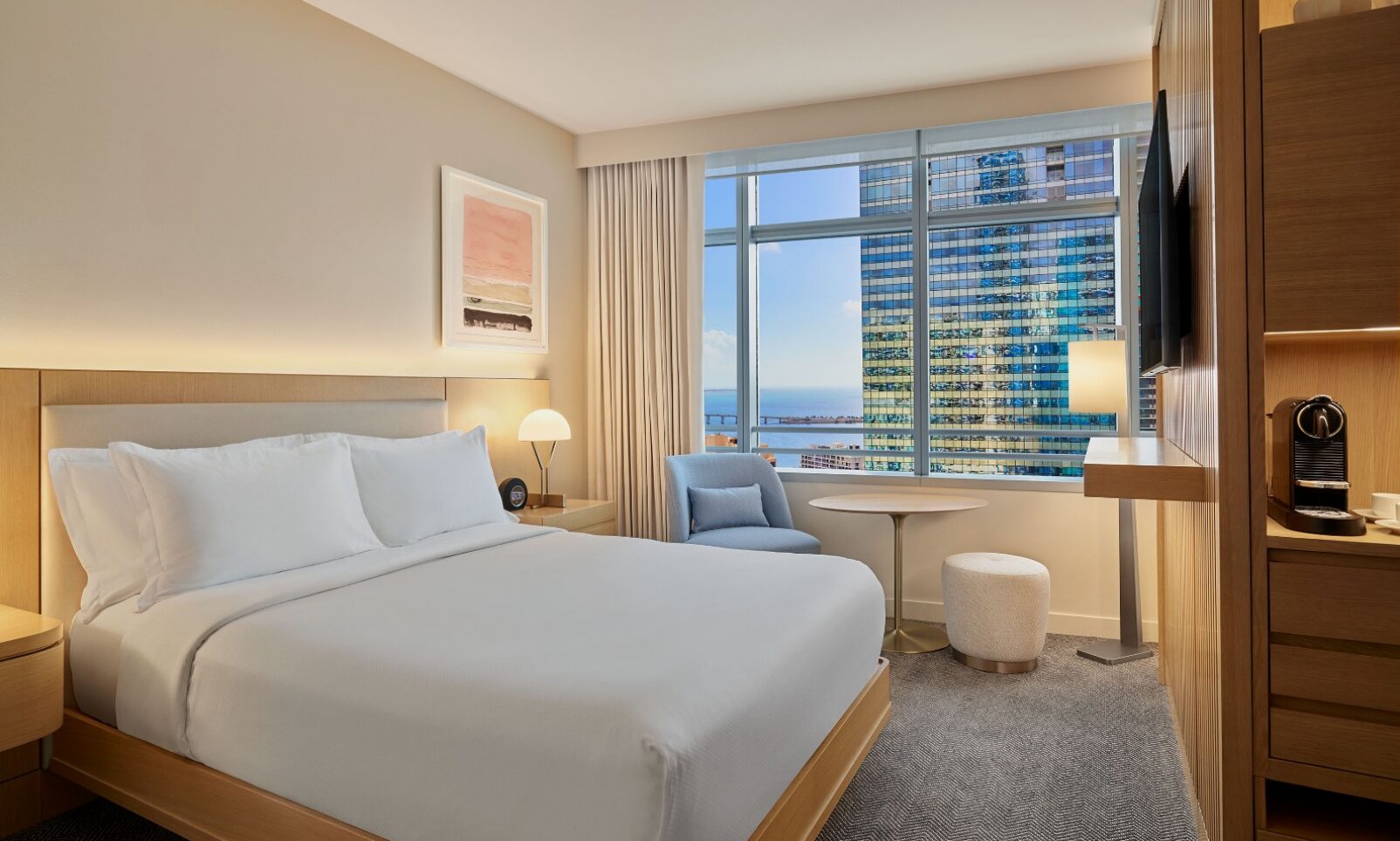 Modern hotel room with a large bed, armchair, desk, TV, coffee maker, and a window offering a city and ocean view.
