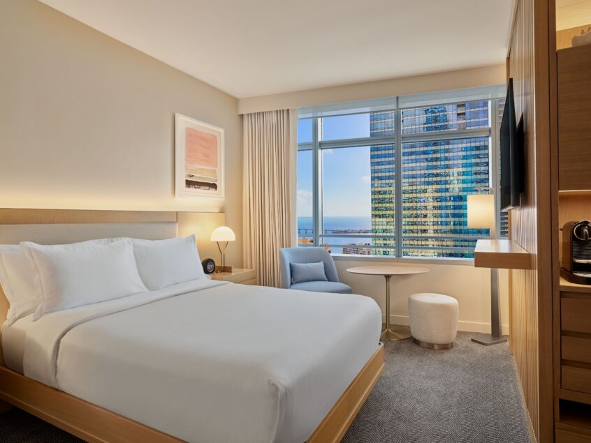 Modern hotel room with a large bed, armchair, desk, TV, coffee maker, and a window offering a city and ocean view.