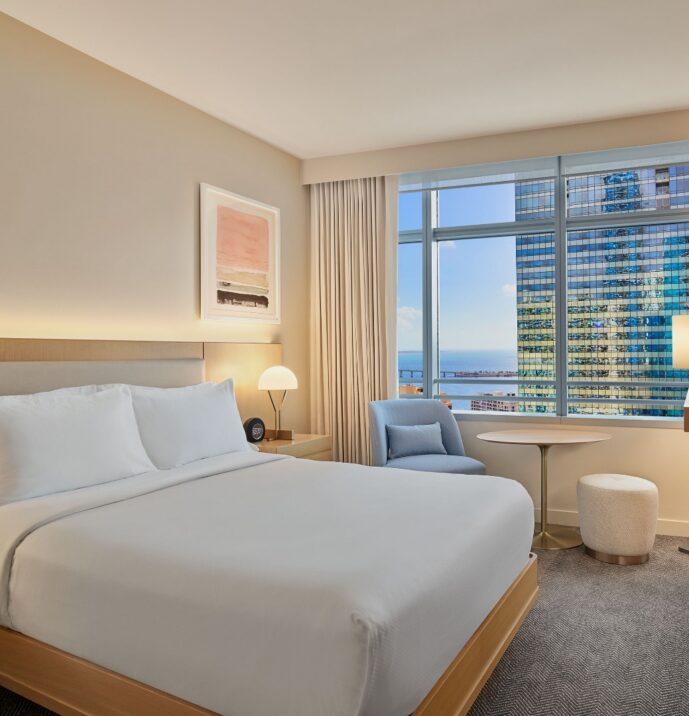 Modern hotel room with a neatly made bed, cozy armchair by a window, city view outside, TV on the wall, and wooden furniture and décor.