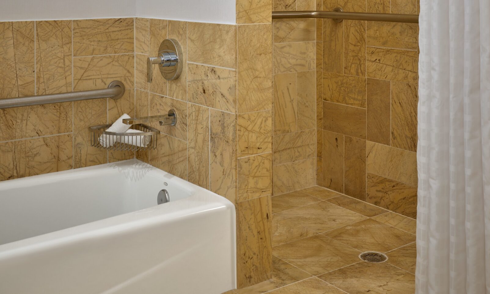 Accessible bathroom with tan stone tiles, a bathtub, a walk-in shower with grab bars, and a white shower curtain.