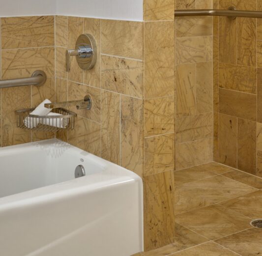 Accessible bathroom with tan stone tiles, a bathtub, a walk-in shower with grab bars, and a white shower curtain.