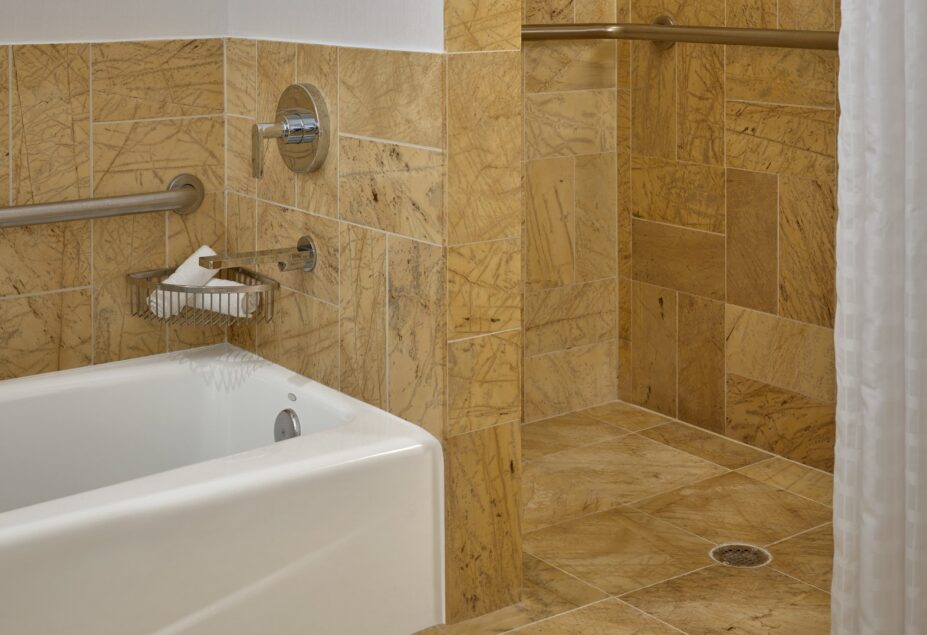 Accessible bathroom with tan stone tiles, a bathtub, a walk-in shower with grab bars, and a white shower curtain.