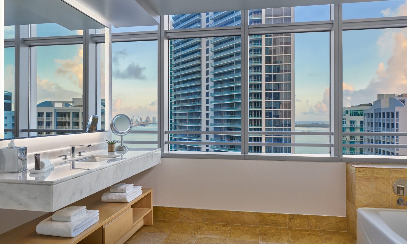 Modern bathroom with large windows, city and water views, a marble vanity, towels on open shelves, and a bathtub beside the window.