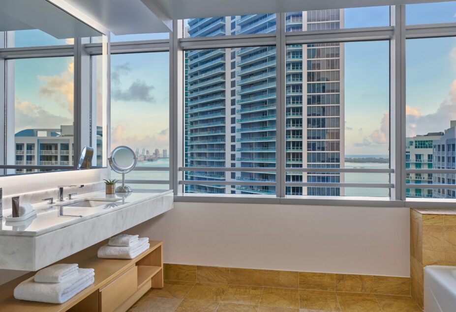 Modern bathroom with large windows, city and water views, a marble vanity, towels on open shelves, and a bathtub beside the window.
