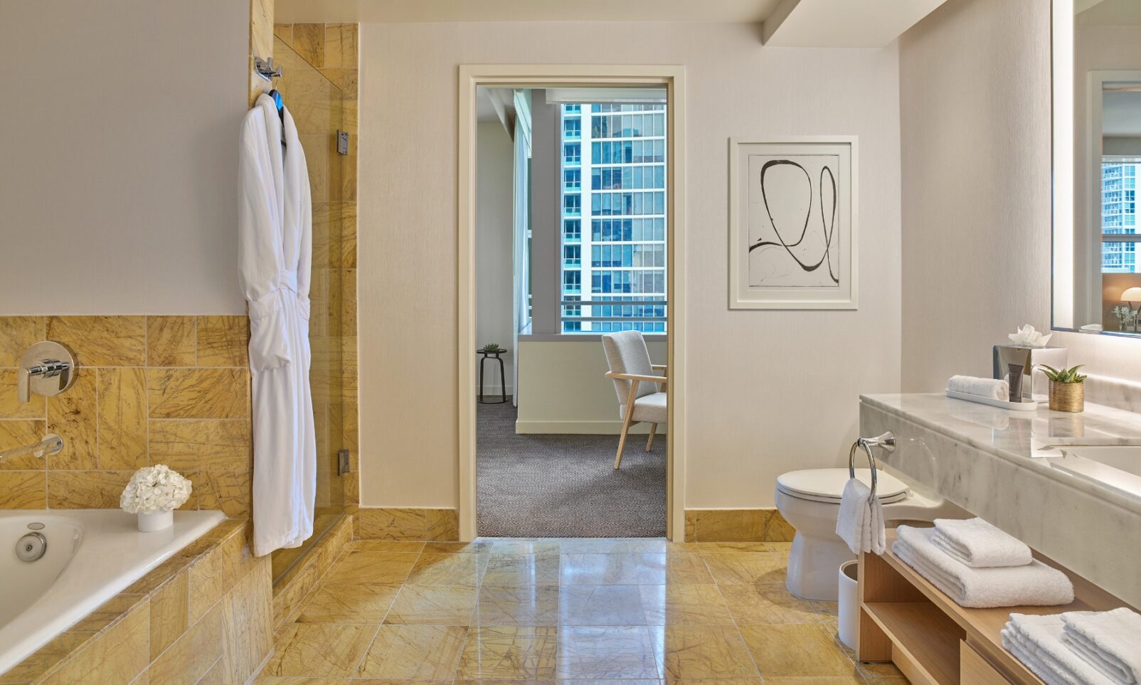 Modern bathroom with tan tiles, a bathtub, white robe, vanity with sink, and a view into a bright room with a chair and city buildings outside.