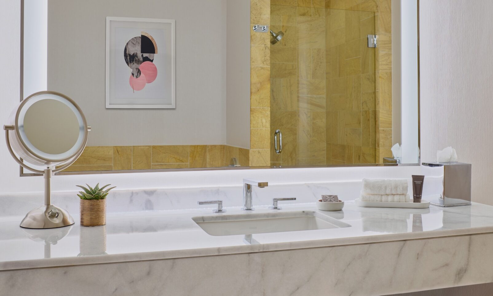 Modern bathroom with marble countertop, sink, mirror, toiletries, small plant, and a walk-in shower with yellow tiles in the background.
