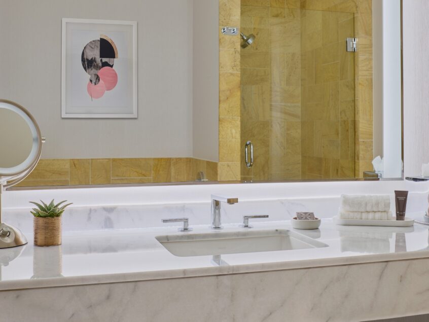 Modern bathroom with marble countertop, sink, mirror, toiletries, small plant, and a walk-in shower with yellow tiles in the background.