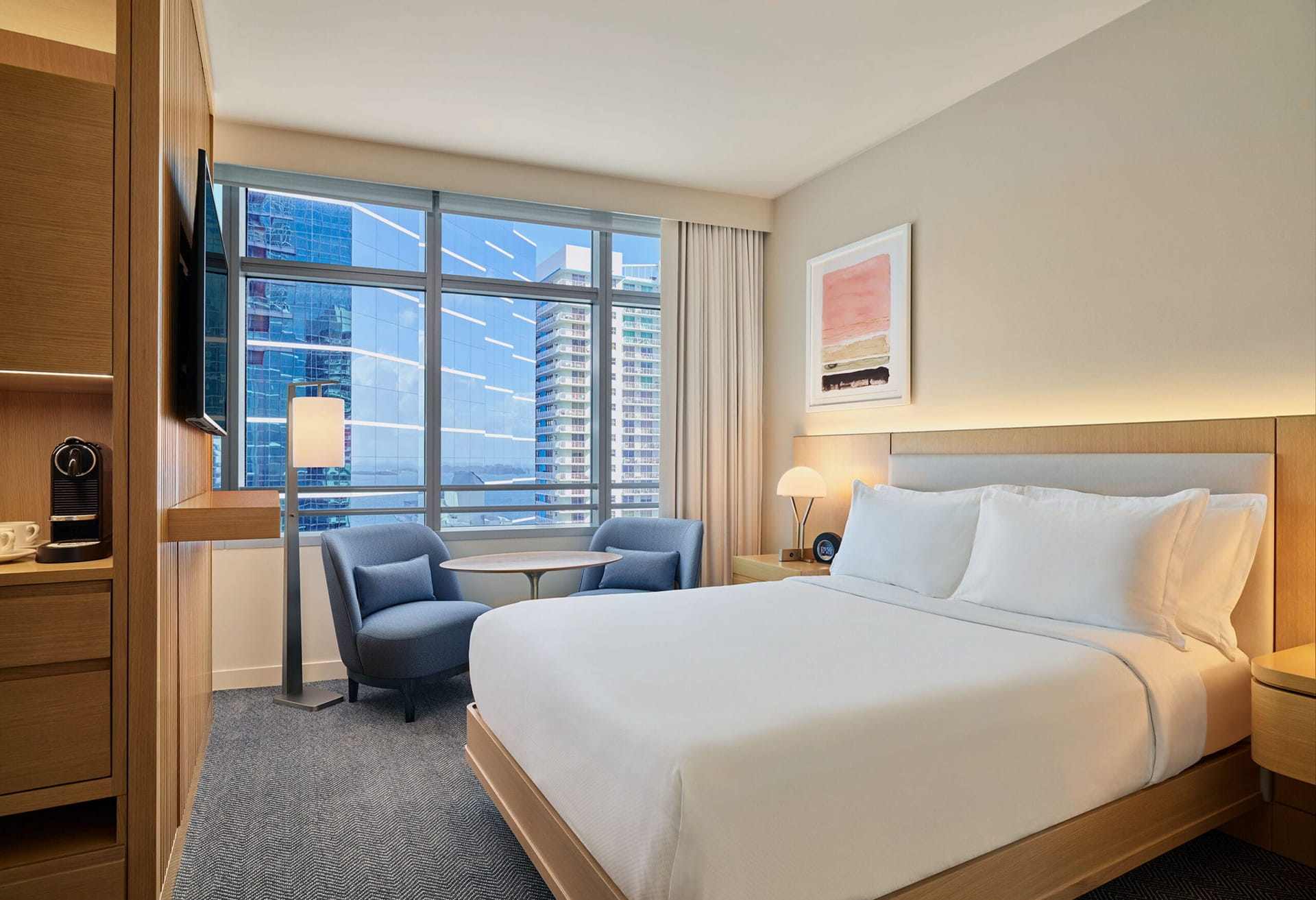 Modern hotel room with a large bed, two blue chairs by a window, a small table, wall art, and cityscape view outside.