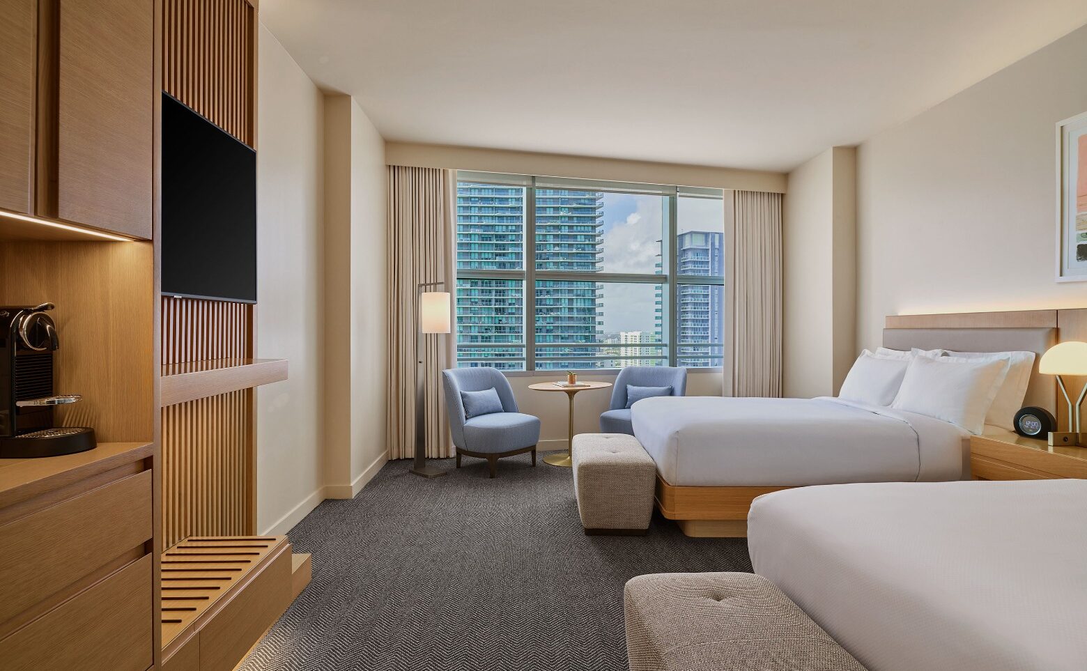 Modern hotel room with two beds, a TV, coffee maker, seating area by a large window, and a cityscape view in the background.