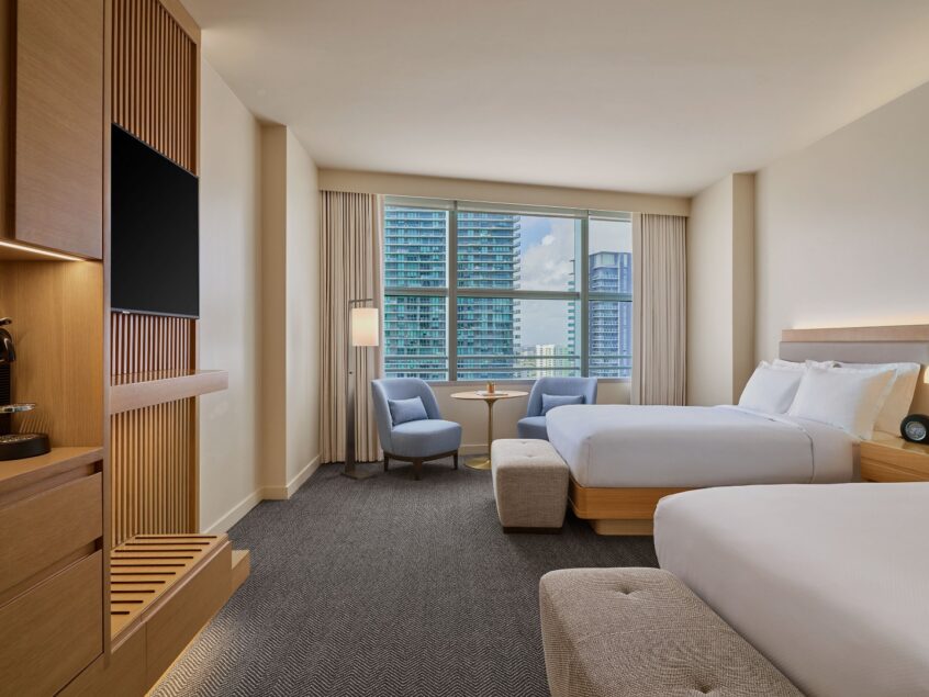 Modern hotel room with two beds, a TV, coffee maker, seating area by a large window, and a cityscape view in the background.