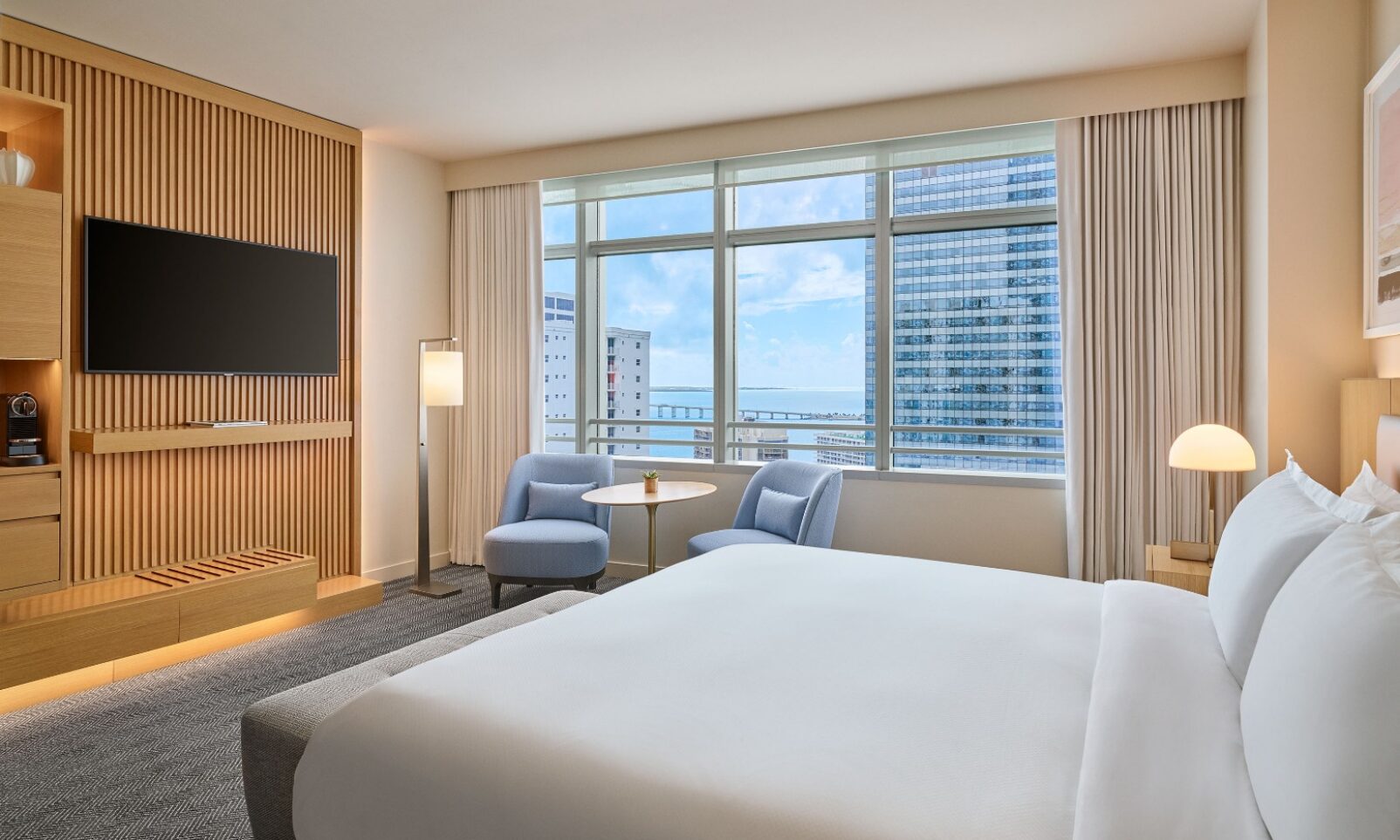 Modern hotel room with a large bed, seating area by a window, city view, wall-mounted TV, and light wood furnishings.