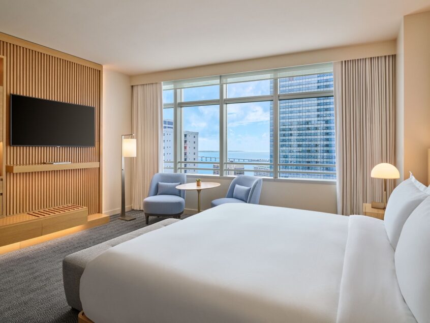 Modern hotel room with a large bed, seating area by a window, city view, wall-mounted TV, and light wood furnishings.