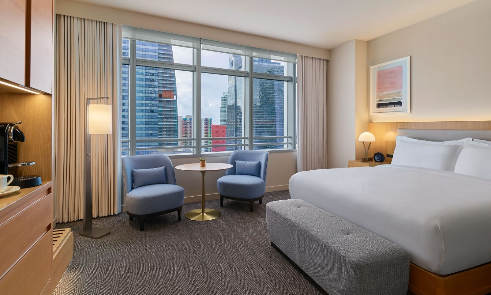 Modern hotel room with a large bed, two blue chairs, a small table, and floor-to-ceiling windows overlooking city skyscrapers.