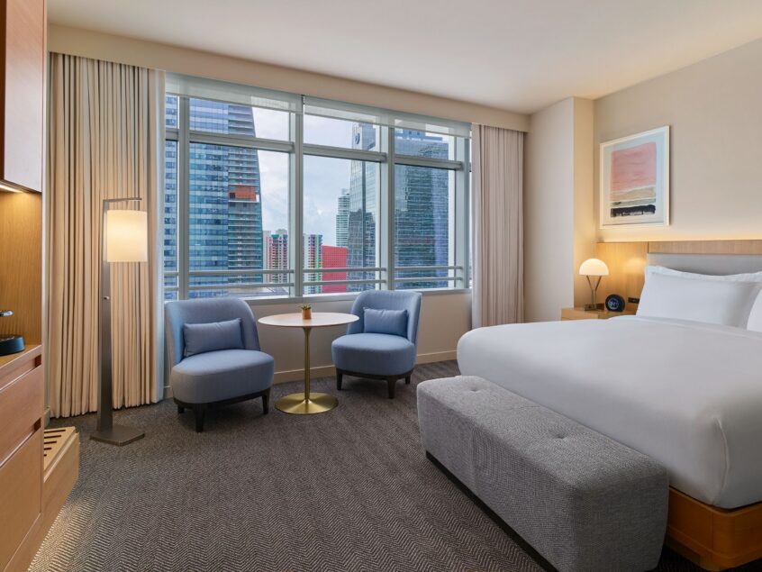 Modern hotel room with a large bed, two blue chairs, a small table, and floor-to-ceiling windows overlooking city skyscrapers.