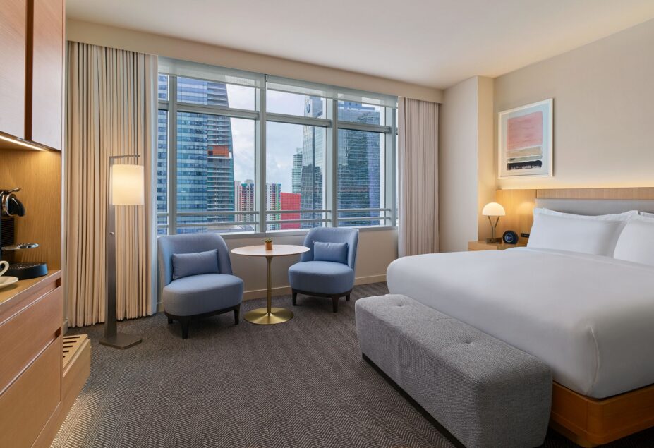 Modern hotel room with a large bed, two blue chairs, a small table, and floor-to-ceiling windows overlooking city skyscrapers.