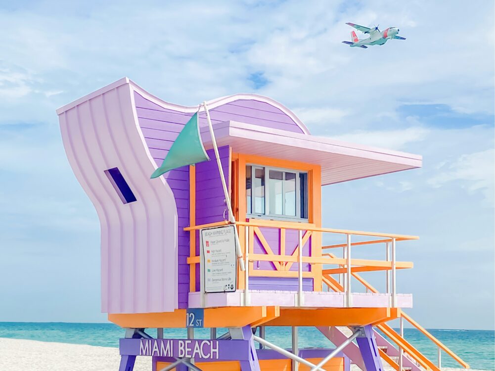A colorful lifeguard tower on Miami Beach with pastel purple and orange accents, under a blue sky with a plane flying above.