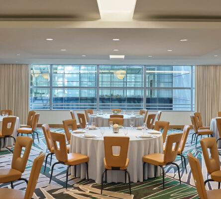 Modern banquet room with round tables set for dining, tan chairs, white tablecloths, large windows, and geometric-patterned carpet.