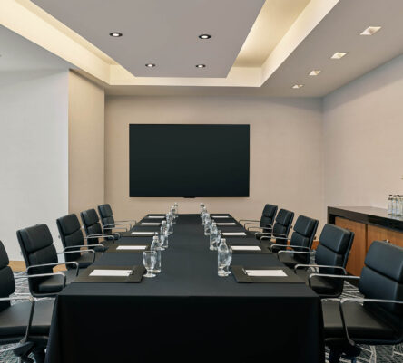 Modern conference room with a rectangular table, black chairs, notepads, water glasses, a blank screen, and a sideboard with flowers and bottled water.