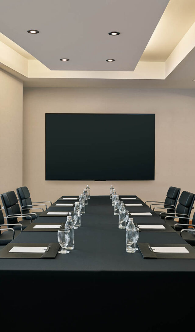 Modern conference room with a rectangular table, black chairs, notepads, water glasses, a blank screen, and a sideboard with flowers and bottled water.