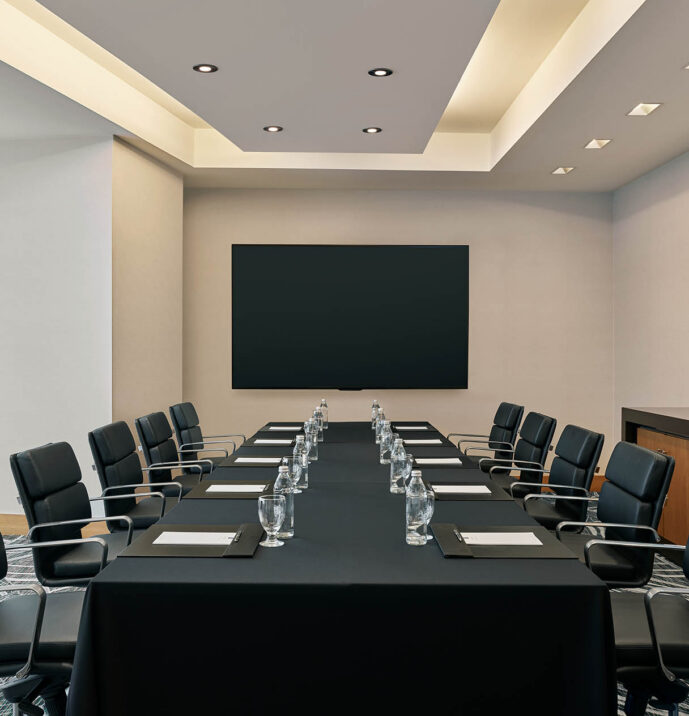 Modern conference room with a rectangular table, black chairs, notepads, water glasses, a blank screen, and a sideboard with flowers and bottled water.