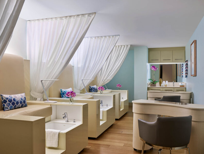 Modern spa room with three pedicure stations, white curtains, plush chairs, flower vases, and soft lighting, creating a relaxing atmosphere.