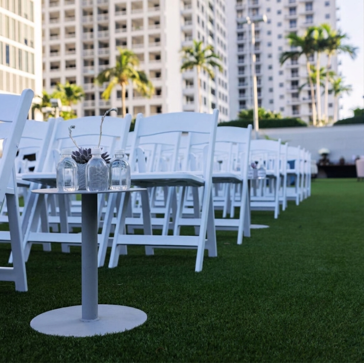 Rows of white folding chairs and a small round table with glass vases on green grass, set outdoors near tall buildings and palm trees.