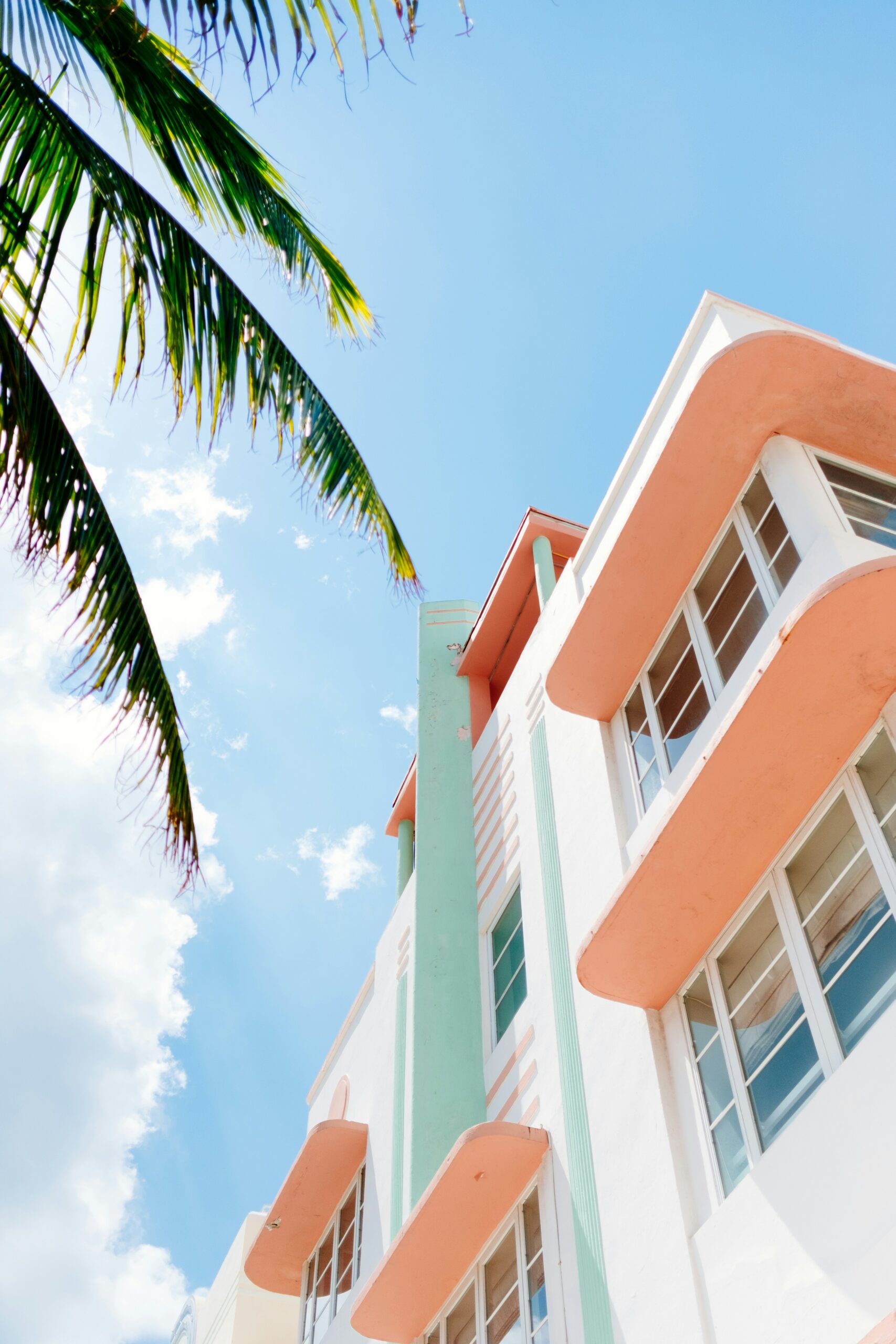 Art Deco building with pastel colors and rounded balconies under a bright blue sky, with a palm leaf in the corner.