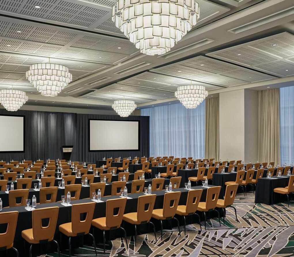 Spacious conference room with rows of tables and chairs, two large screens, chandeliers, and floor-to-ceiling windows with sheer curtains.