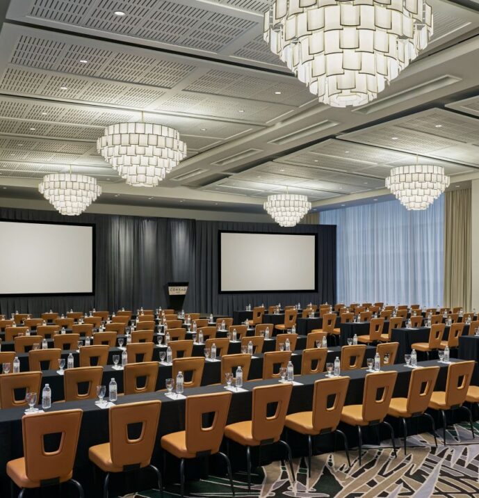 Spacious conference room with rows of tables and chairs, two large screens, chandeliers, and floor-to-ceiling windows with sheer curtains.