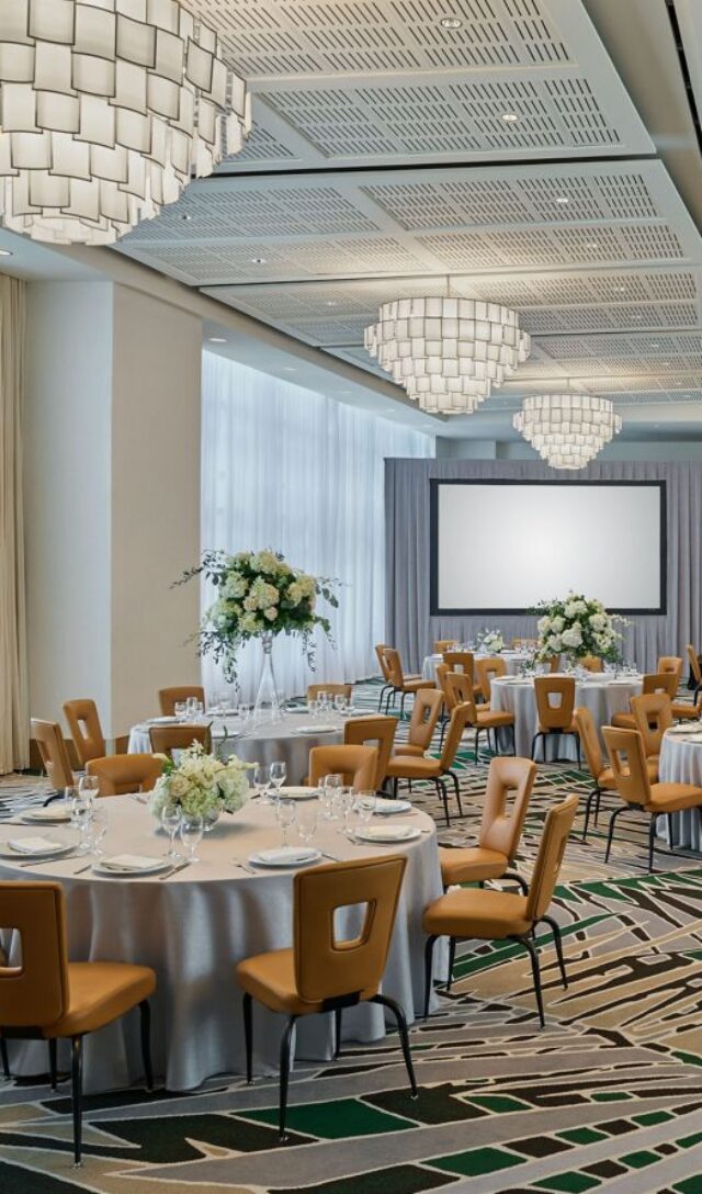Elegant banquet hall with round tables, tan chairs, floral centerpieces, large chandeliers, and two projection screens at the front.