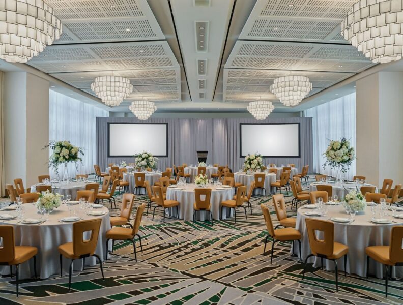 Elegant banquet hall with round tables, tan chairs, floral centerpieces, large chandeliers, and two projection screens at the front.