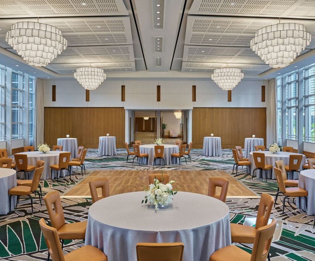 Spacious banquet hall with round tables, brown chairs, tall windows, modern chandeliers, and white tablecloths set for an event.