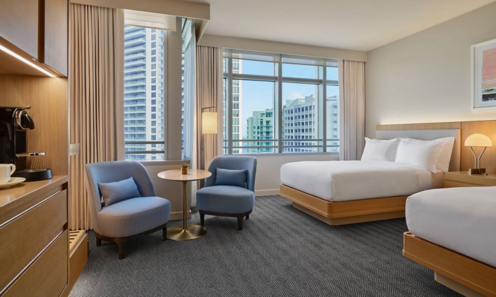 Modern hotel room with two double beds, light wood furniture, blue chairs around a small table, large windows, and city buildings visible outside.