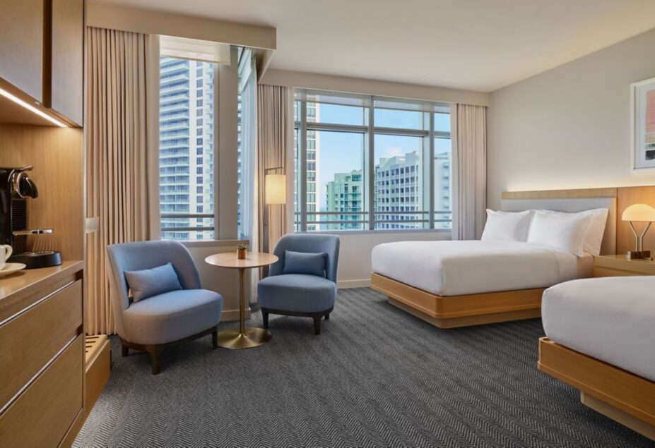 Modern hotel room with two double beds, light wood furniture, blue chairs around a small table, large windows, and city buildings visible outside.