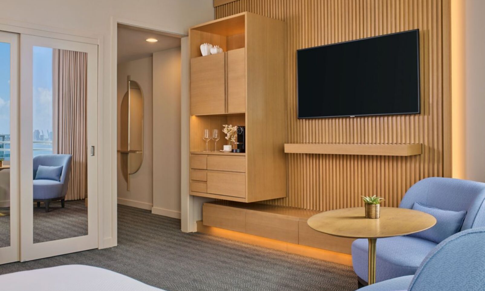 Modern hotel room with a wall-mounted TV, wooden cabinetry, blue chairs, a round table, and large sliding glass doors leading to a bright sitting area.