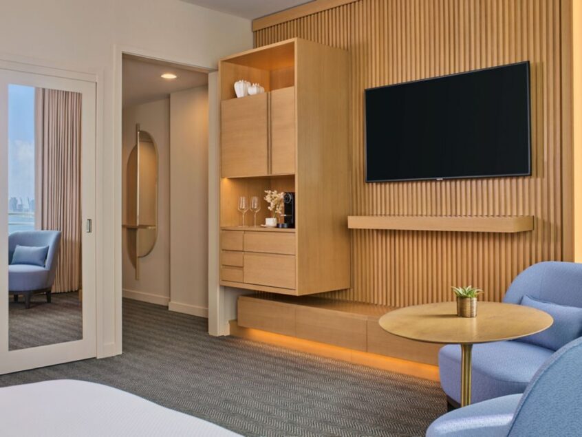 Modern hotel room with a wall-mounted TV, wooden cabinetry, blue chairs, a round table, and large sliding glass doors leading to a bright sitting area.